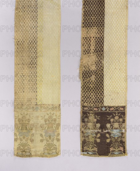 Kontusz belt, flower bouquets ornament, kontusz belt men's national costume accessory, Poland, carp scale ornament, bipartite heads with a floral bouquet pattern on a base, colors: ochre, blue, green, brown, yellow, dated 1768–1780, place of origin Kutkorz, clothing accessory, length 290 cm, width 29 cm, A close-up of two long vertical textile strips laid side by side on a pale background, each strip showing decorative woven patterns and areas of wear; the left strip features a pale creamy yellow panel and a darker golden-beige patterned panel with a repeating scallop motif, and a lower section showing a floral border with muted green and pale blue accents on a light yellow-beige ground; the right strip shows a dark brown patterned panel with a repeating scallop motif alongside a pale yellow panel, and a lower floral border with faded green and pale blue details on a dark brown ground, with some fraying and worn patches revealing lighter threads.