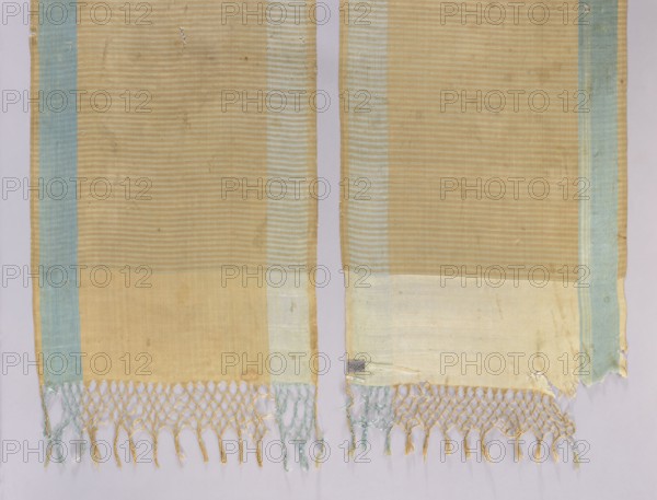 Kontusz belt, Polish kontusz belt men's clothing accessory, national costume from 18th-century Poland, associated with Szydlów Swietokrzyskie Voivodeship. Smooth heads; narrow, smooth two-tone stripes; edge made of a wide smooth belt. Silk fringe, partially braided. Colors: crimson, blue, and white. Dimensions: length 300 cm, width 47 cm., A woven textile shown in two vertical panels with a background surface, each panel primarily beige with subtle horizontal stripe texture, bordered on one side by a pale blue stripe and on the other by a very light blue or off-white stripe; the lower portions of both panels have a wider pale beige or cream band. The bottom edges have a knotted fringe with twisted tassels in a mix of beige and pale blue threads, and some fraying and small holes appear along the fabric edges. The overall colors visible are beige, pale blue, off-white, and hints of light tan on a light neutral background.