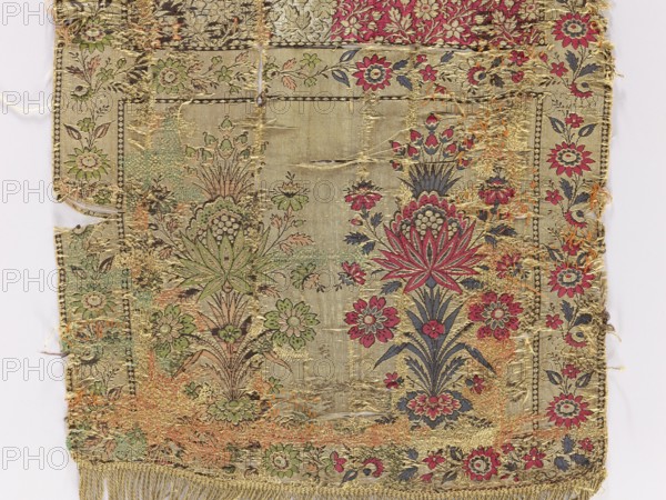 Kontusz belt, Polish clothing accessory. 2nd half of the 18th century, Lyon, France. Four-sided, bipartite heads with a floral bouquet pattern; continuous pattern of flower branches with tendrils of large flowers. Sewn-on gold fringe. Materials/colors: gold, light green, pink, navy blue, cherry red, black. Length 174 cm, width 36 cm., A worn rectangular textile fragment with a floral pattern, showing an ornate central panel flanked by decorative borders; the fabric background is a beige or light gold tone with visible areas of fraying and thread loss. The design includes stylized flowers and foliage in shades of red, pink, green, dark blue, and muted brown, with gold or yellow metallic threads woven throughout; some floral motifs feature a prominent starburst-like bloom in red and dark blue, and other blossoms in green and pink. The borders contain repeating floral sprigs in similar colors, and the lower edge has a short fringe in a yellowish-gold color. There are patches where threads...