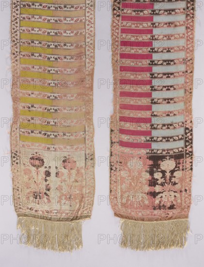 Silk kontusz sash, four-sided, c. 1787–1794. Likely made by Szczepan Filsjean Manufactory Kobylka. Bipartite ends each with a bouquet motif of rooted carnations tied with a ribbon. Field in transverse stripes alternating plain or two-coloured with fine floral tendrils; trim with small flowers. Borders vertical and horizontal with twig motifs and checkerboard. Sewn-on silk fringe. Colors: cream, brown, yellow-green, sky blue, dark red; right side: cream background of heads, borders and shelves with tendril, brown ornament; plain shelves half pink, half yellow-green; reverse: plain shelves half sky blue, half dark red. Weave density approx. 17/39 x2. Length 357 cm, width 32 cm. Origin: Kobylka, Masovian Voivodeship, Poland., Two long narrow textile panels hang side by side, each showing horizontal bands and floral motifs with fringed ends. The left panel features alternating horizontal stripes in shades of pale yellow and beige with narrow decorative bands in muted pink and dark brown, a...