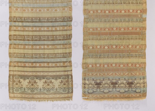 Kontusz belt. Quadripartite heads with a pattern of flower bouquets in inverted vases; earth marked at the bottom and clouds at the top. Alternating transverse stripes, smooth and varied in color, with tendril motifs in three varieties; partial edging with plant motifs. Colors: beige, brown, light blue, cream, pink, green. Reversible belt, men's clothing accessory. 2nd half of the 18th century, Volhynia. Length 290.5 cm; width 44.5 cm., Two long woven textiles are visible side by side against a pale background, each showing horizontal patterned bands with repeating motifs. The left textile has a muted palette of beige, light yellow, pale blue, soft brown, and faded green, with decorative floral and scroll-like designs and a fringe at the lower edge. The right textile features similar horizontal banding with colors including beige, warm tan, muted orange, pale blue, brown, and green, also showing floral and ornamental patterns and a slightly frayed lower edge. Both pieces display altern...
