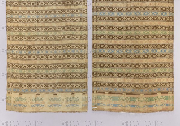 Kontusz belt, One-sided men's kontusz belt with transverse stripes featuring stylized plant tendrils and geometric ornamentation. The belt's head ornament consists of four reversed floral patterns; the frame features stylized floral and geometric ornamentation. Predominant colors: sand, light green, blue-pink, and brown. 18th century, possibly Russia. Length 320 cm, width 55 cm., The image shows a pair of hanging rectangular textile panels with repeating horizontal bands of ornamental floral and geometric motifs. The fabric displays a palette of beige and tan as the dominant background color, with darker brown used for many of the patterned stripes. Accents of muted blue and soft green appear in several bands and in larger stylized floral motifs near the lower edges. The lower borders of both panels feature larger decorative floral shapes in pale blue and green against the beige ground. The panels are separated by a narrow vertical gap revealing a pale neutral background, and the botto...