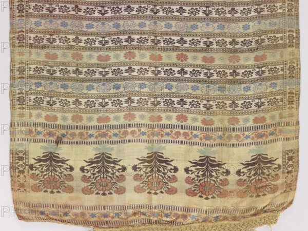 Kontusz sash; Five-part heads with an ornament of stylized, inverted floral bouquets alternating with transverse stripes bearing small Persian palmettes, floral tendrils, and a fine wavy pattern. Silk fringe. Predominant colors: light yellow, brick red, brown, and sapphire blue. 18th century, Russia; national costume accessory. Length 331 cm, width 64.5 cm. Materials: silk., A woven textile with horizontal bands of repeating floral and geometric motifs in colors including beige, brown, blue, green, and muted orange; the top and middle sections show several narrow stripes filled with alternating flower shapes and small leaf-like patterns in brown and blue on a beige background; below these are a few bands with larger floral clusters in brown and muted orange set against pale greenish-beige fields; near the bottom is a wider area with repeated stylized tree-like floral arrangements in dark brown with orange flower centers on a light beige ground, and the lower edge has a decorative woven...
