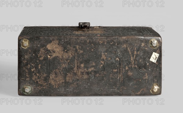 Casket, plant decoration, shields, dates, geometric ornament, early 16th century, possibly France, Flanders, or Germany; piece of furniture; height 12 cm, width 26.5 cm, depth 12.5 cm., A rectangular box shown from the front and top with a weathered dark brown to black surface and worn lighter brown patches, featuring incised decorative lines and carved star-like shapes; the box has a hinged clasp on top and rounded corners with several circular metal fittings that appear brass or bronze with greenish patina; a small light-colored rectangular paper tag is attached near one corner; the background is a neutral light gray.
