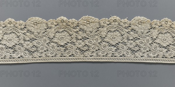 Lace fragment, machine-made lace, strip serrated on one side in accordance with the forms of rosettes and flower cups placed on the edge, late 19th century, Europe, width 9.5 cm, length 100 cm., A horizontal band of intricate lace with scalloped upper edge stretches across a flat background, showing a repeating floral and circular openwork pattern with delicate loops and connecting threads; the lace is a cream or off-white color and the background is a muted gray, with several small shadowed openings visible within the lace and fine linear edging along the bottom.