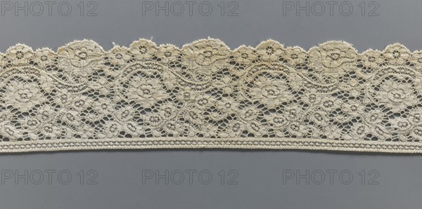 Lace fragment, machine-made lace; strip serrated on one side following the forms of rosettes and flower cups placed on the edge. Late 19th century, Europe. Lace; width 9.5 cm, length 100 cm., A horizontal piece of lace with an intricate openwork pattern rests against a flat background; the lace is a pale beige color with repeating floral and scroll motifs and scalloped upper edges, showing delicate woven loops, small circular flower shapes, and interconnected net-like openings, while the background is a solid medium gray color.