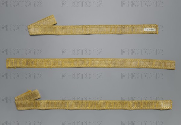Three one-sided galloons; creator, galloons haberdashery; origin, 18th century, Poland; fabrics; width 3.2 cm, length 100 cm., A photograph shows a neutral gray background with a few long, narrow woven bands laid out horizontally across it. The bands are primarily gold in color with areas of lighter and darker gold tones, and they have decorative woven edges and textured interior patterns. One band at the top has a folded end, and the bottom band also shows a folded segment, while the middle band lies flat. The bands display slight fraying and uneven edges in places, and the overall scene contains shades of gray from the background and various shades of gold on the woven pieces.