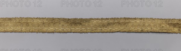 Five gallons, gallons haberdashery, origin unknown, geometric ornaments, 18th century, Europe, fabrics, width 1.5 cm, length 63 cm., A long narrow woven band lies horizontally across a plain light gray background, the band showing a metallic golden color with variations in sheen, a few darker bronze-like areas, and tiny hints of silver where threads catch the light; the band has a textured woven pattern with repeating rounded motifs and slightly frayed edges, and the overall palette consists of gold, bronze, silver highlights, and light gray.