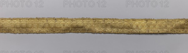 Five gallons, gallons haberdashery, origin unknown, geometric ornaments, 18th century, Europe, fabrics, width 1.5 cm, length 63 cm, A horizontal narrow woven band runs across the center of the image against a smooth background, showing a metallic golden color with subtle variations in tone; the band has a textured, patterned surface made of interlaced threads that create repeating oval and linear motifs, and its edges are slightly irregular with tiny loose fibers; the background is a uniform pale gray that contrasts with the warm gold of the band.