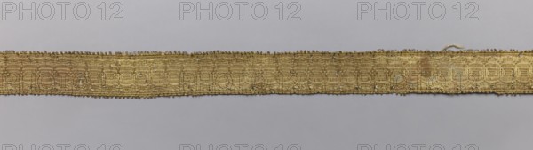 Five gallons, gallons haberdashery, origin unknown, geometric ornaments, 18th century, Europe, fabrics, width 1.5 cm, length 63 cm., A narrow woven band runs horizontally across the image against a pale gray background, showing a textured surface of gold threads with a subtle sheen and small irregularities along its edges; the band displays a repeating woven pattern with areas of slightly darker gold and hints of lighter, almost silvery highlights, and a few tiny loose fibers and small gaps are visible along its length.