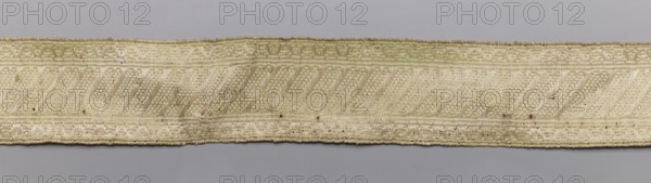 Three gallons, gallons haberdashery, origin unknown, 19th century, Europe, fabrics, width 3.5 cm, length 76 cm., A narrow woven band stretches horizontally across a gray background, showing a textured surface with a repeating diagonal rib pattern and decorative borders; the band is mostly beige and off-white with areas of pale yellowing and a few small brown spots and speckles, and its edges appear slightly frayed.