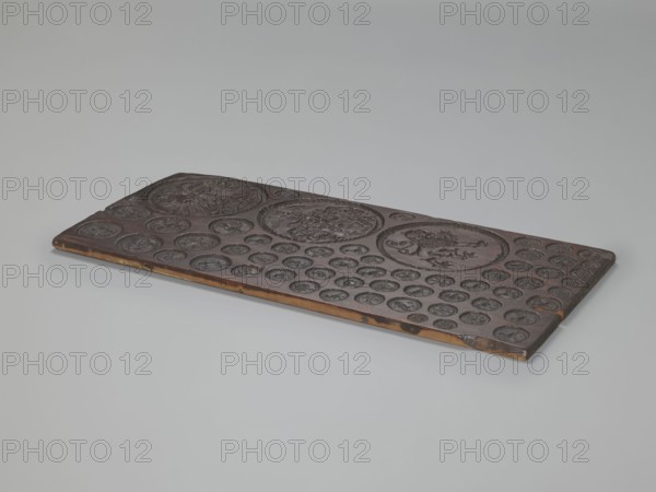 Gingerbread mold with 3 coats of arms and 52 medallions, boards and gingerbread, national emblems, zoomorphic motifs, coats of arms, banners, female busts, decorative art. Rectangular, flat gingerbread mold with representations that form medallions of various sizes, with a wreath-shaped border. Three large medallions, placed one below the other, contain heraldic images: 1. A double-headed eagle under a crown. 2. A quadripartite coat of arms, in whose fields appear a lion, an eagle, a human figure, and the letter W; in the central field a human head; the coat of arms topped with a crown, above which are a female bust and two banners; on the sides of the shield are decorative labres made of plant twigs. 3. A mounted lion facing right. The form's decoration is complemented by 52 small medallions containing images of animals mostly birds, and anthropomorphic, geometric, and plant motifs. 16th–17th century, utility item., A flat rectangular wooden panel with a dark brown surface and lighter...
