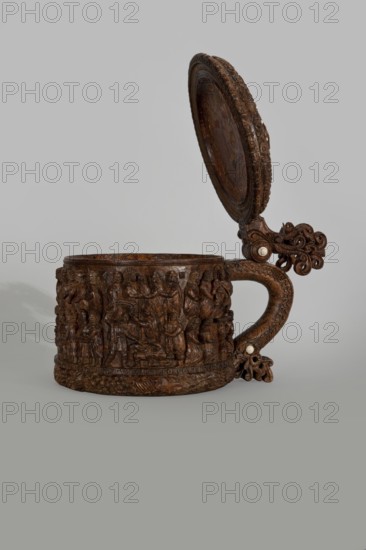 Wooden pear-shaped peg tankard, round, richly decorated with bas-reliefs depicting Passion scenes and a Resurrection of Christ scene on the lid; lid rim and handle decorated with plant motifs and masks and studded with ivory pins; center of lid has a row of vertically driven metal pegs placed at regular intervals top two missing. Carved wood, inscriptions, figurative and Gospel/Passion iconography, noted. 17th century, Scandinavia. Height 23.5 cm; width 20.5 cm., An ornately carved wooden tankard with a hinged lid is shown against a plain gray background; the body and handle display intricate relief scenes and scrolling decoration, with the lid opened upright to reveal detailed carved work along its rim and inner surface, and there are a few small pale inlaid circular accents on the hinge and handle; the visible colors are brown tones in the wood, pale cream in the inlays, and gray in the background.