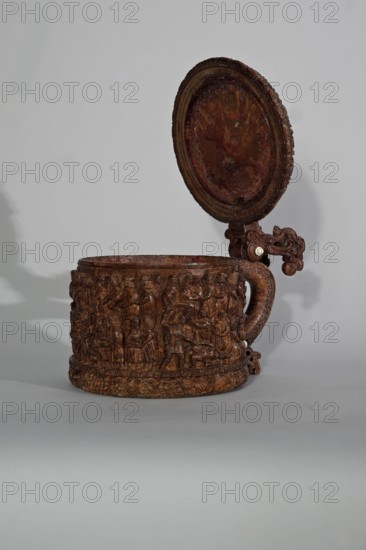 Wooden tankard, carved, probably a peg tankard, religious scenes, Gospel scenes, Resurrection of Christ iconography, inscriptions, vessels, Passion Bible; New Testament, human figures, figurative motifs, decorative art. Wooden pear-shaped tankard, so-called peg tankard, round, richly decorated with bas-reliefs depicting Passion scenes, with a Resurrection scene on the lid. The lid rim and handle are decorated with plant motifs and masks, and studded with ivory pins. In the center is a row of vertically driven metal pegs placed at regular intervals the top two are missing. 17th century, Scandinavia. Vessel. Height 23.5 cm, width 20.5 cm., A carved wooden tankard with an open lid sits against a plain light gray background; the tankard is a deep brown color with darker and lighter variations, and its cylindrical body is covered in intricate relief carvings depicting a group of human figures and decorative patterns, while the hinged circular lid shows worn textures and circular carving det...