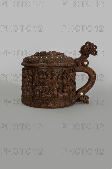 Wooden pear-shaped tankard, so-called peg tankard, carved, round, richly decorated with bas-reliefs depicting Passion scenes and a Resurrection of Christ scene on the lid; lid rim and handle decorated with plant motifs and masks and studded with ivory pins; center has a row of vertically driven metal pegs placed at regular intervals top two missing. Inscribed; figurative motifs of Gospel/Passion scenes and human figures. 17th century, Scandinavia. Vessel. Height 23.5 cm, width 20.5 cm., An intricately carved wooden vessel with a domed lid and a curved handle sits against a plain neutral background; the vessel displays detailed relief scenes around its cylindrical body and ornate scrolling decorations on the handle and lid, with small round insets visible on the lid and handle; the object is predominantly brown with lighter ivory-colored round accents and subtle variations in wood tone, set on a gray backdrop.