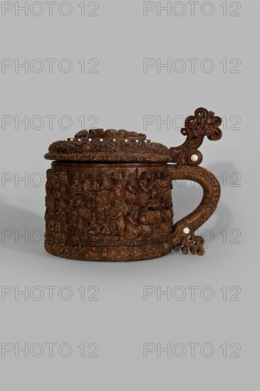 Wooden tankard, carved, probably a peg tankard;, Pear-shaped wooden tankard, round, richly decorated with bas-reliefs depicting Passion scenes, with a Resurrection of Christ scene on the lid. Lid rim and handle decorated with plant motifs and masks, and studded with ivory pins. Center of the lid has a row of vertically driven metal pegs placed at regular intervals the top two are missing. Inscriptions present. Figurative motifs and human figures; Gospel/Passion iconography. Function: vessel; decorative art; . 17th century, Scandinavia. Height 23.5 cm, width 20.5 cm., A carved wooden tankard with a hinged lid and a curved handle sits against a light grey background; the tankard is dark brown with carved relief scenes wrapping its body and lid, and it is adorned with a few small round light-colored insets on the lid and handle; the handle features ornate scrollwork and decorative flourishes, and the surface shows textured, detailed carving throughout.