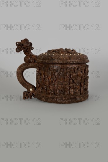 Wooden pear-shaped peg tankard, carved, round, richly decorated with bas-reliefs depicting Passion and Gospel scenes, with a Resurrection of Christ scene on the lid; lid rim and handle decorated with plant motifs and masks and studded with ivory pins; center of lid has a row of vertically driven metal pegs placed at regular intervals top two missing. Inscribed; human figures and figurative motifs., noted. Decorative art; vessel. 17th century, Scandinavia. Height 23.5 cm, width 20.5 cm., A carved wooden lidded vessel with a handle is shown against a plain gray background; the vessel is dark brown with a slightly glossy finish and has a richly detailed relief band around its body depicting many small human figures and scenes, and the lid is adorned with carved decorative motifs and a ring of small light-colored round inlays. The handle curves outward and features ornate scrollwork with a carved flourish at its end and a couple of small pale inlays near its base; the base of the vessel ha...