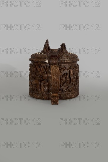 Wooden pear-shaped tankard, probably a peg tankard, carved, round, richly decorated with bas-reliefs depicting Passion scenes, with a Resurrection of Christ scene on the lid; lid rim and handle decorated with plant motifs and masks and studded with ivory pins; center has a row of vertically driven metal pegs placed at regular intervals the top two are missing. Inscriptions; religious/Gospel motifs; human figures; decorative art;, noted. 17th century, Scandinavia. Vessel; height 23.5 cm, width 20.5 cm., A carved round wooden container with a lid sits on a smooth surface against a light gray background; the object is dark brown with areas of lighter brown highlights and small pale dots along the lid edge, and it features intricate raised carvings of figures and decorative patterns around its sides, a prominent carved vertical handle or strap on the front with detailed motifs, and sculpted elements on top that rise above the lid.