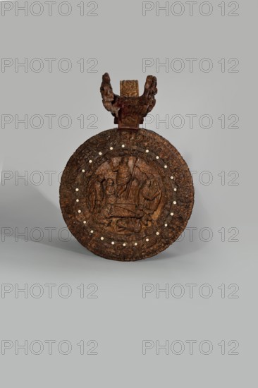 Wooden tankard, carved, probably a peg tankard, Depicts religious/Gospel scenes, including Passion scenes in bas-relief and a Resurrection of Christ on the lid. Inscriptions present. Pear-shaped wooden tankard, round, richly decorated with bas-reliefs of Passion scenes; lid rim and handle decorated with plant motifs and masks, studded with ivory pins. Center has a row of vertically driven metal pegs placed at regular intervals top two missing. 17th century, Scandinavia. Vessel; height 23.5 cm, width 20.5 cm., A carved circular wooden object with a carved decorative handle at the top, shown against a plain light gray background. The object is predominantly dark brown with carved figures and textured patterns across its surface, and a ring of small round light-colored inlaid spots around the central carved area. The handle has ornate carved elements in dark brown matching the circular piece, and the lower shadow indicates it is resting on a flat surface.