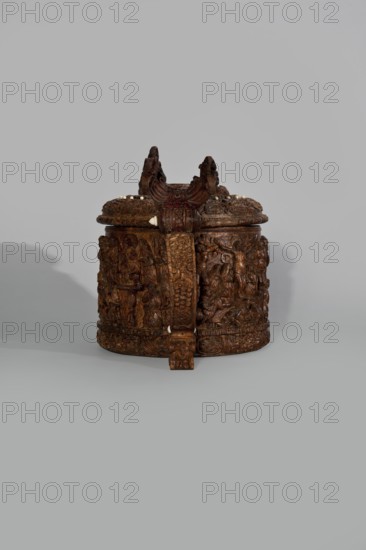 Wooden tankard, carved, probably a peg tankard, Religious/Gospel scenes: Passion scenes in bas-relief around the body and a Resurrection of Christ scene on the lid. Inscriptions; human figures and figurative motifs., noted. Decorative art: wooden pear-shaped tankard, round, richly decorated; lid rim and handle decorated with plant motifs and masks, studded with ivory pins. Center has a row of vertically driven metal pegs placed at regular intervals the top two missing. 17th century, Scandinavia. Vessel. Height 23.5 cm; width 20.5 cm., An ornately carved round wooden container with a lid, showing a dark brown wood tone with lighter brown highlights and small white inlays, decorated with detailed relief scenes and figures around its sides and lid, featuring a prominent vertical carved strap and a raised decorative crest on top, all set against a plain light gray background with a soft shadow to one side.