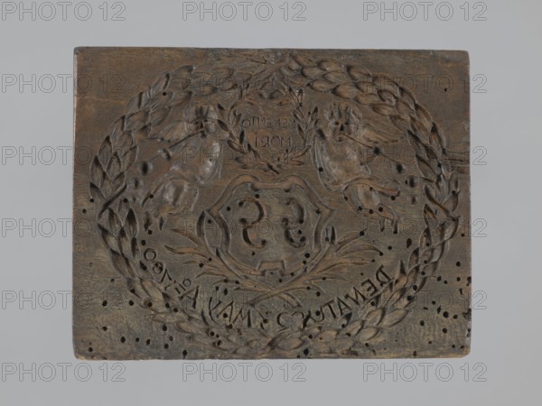 Double-sided gingerbread mold. Obverse: coat of arms with initials GG, top inscription Memento mori, two cherubs at the sides, bottom inscription DENATUS 23 May A 1700, laurel wreath. Reverse: intertwined monogram CMP under a crown. Decorative crown decorated with geometric and plant ornaments. Materials/function: gingerbread, applied/decorative art; motifs: wreath, Latin aphorisms, knotted monograms, plant ornaments, cartouches, angel with trumpet, epitaphs. Date: 1700. Utility item. Dimensions: height 5 cm, width 15 cm, depth 12 cm., A rectangular carved wooden panel with a decorative relief featuring a circular laurel wreath surrounding a central shield-like motif, with two cherub-like figures positioned above the shield and ornamental foliage around them; the wood shows a dark brown tone with lighter brown highlights where carving is worn, and there are several small dark holes scattered across the surface; the background around the panel is a pale gray.