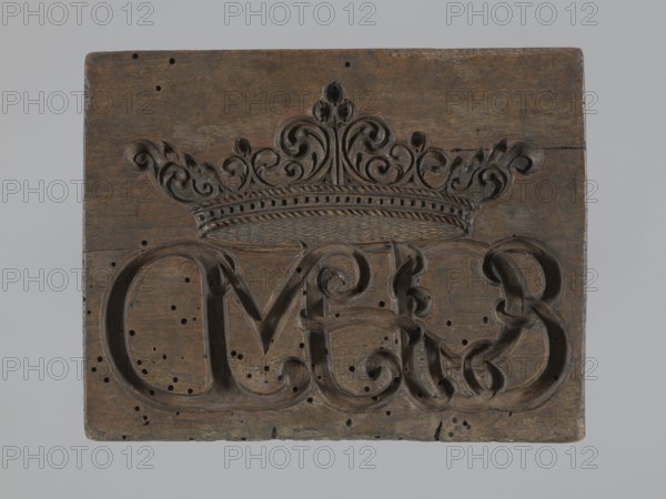 Double-sided gingerbread mold. Obverse: coat of arms with initials GG, at the top the inscription Memento mori, two cherubs at the sides, at the bottom the inscription DENATUS 23 May A 1700 around a laurel wreath. Reverse: intertwined monogram CMP under a crown. Decorative crown decorated with geometric and plant ornaments. Materials/function: gingerbread mold, applied/decorative art, utility item. Motifs: wreath, knotted monograms, plant ornaments, cartouches, angel with trumpet, Latin aphorisms, epitaphs. Date: 1700. Dimensions: height 5 cm, width 15 cm, depth 12 cm. Maker: unknown., A rectangular wooden panel with a carved decorative crown motif at the top and ornate scrollwork below, showing a carved raised design with flowing curves and loops; the wood surface is worn and pitted with a few small round holes and dark spots scattered across it, and the edges are slightly rounded and chipped; visible colors include various shades of brown in the wood, darker brown in the carved reces...