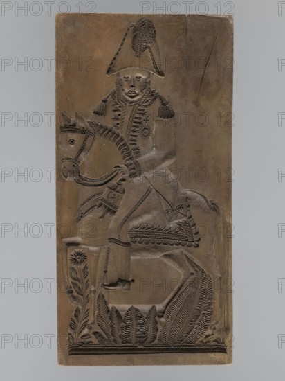 Gingerbread mold, double-sided. Obverse: image of a rider on a horse in a parade uniform from the Napoleonic era 1799–1815, wearing a bicorn, caftan with epaulettes decorated with braiding, an order on the chest, striped trousers and long boots; horse with decorative geometric motif; bottom decoration of leaves and flowers. Reverse: small image of a beehive. Rectangular wooden mold, edge carved with the letters CG Gl. initials. 18th–19th century, Province of Silesia 1742–1918. Height 51.5 cm, width 25.5 cm, depth 5 cm. Possible maker: woodcarver or gingerbread maker; subjects include Napoleonic soldiers, riders, floral decorations, historical military costume; decorative/applied art., A vertical rectangular carved panel showing a figure wearing a tall hat and ornate clothing seated on a horse; the carving is in shades of brown with darker incised lines forming details. The rider has a visible face with carved eyes, nose, and mouth and textured hair or headgear, and the coat features de...