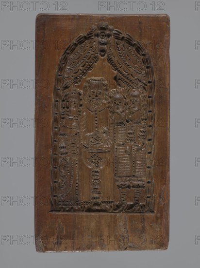 Double-sided gingerbread mould. Obverse: baptism scene; reverse: floral ornament. Genre scenes; gingerbread mould; folk-style decorations; baptisms; floral and plant motifs; applied decorative art; figurative scenes; historical costumes. Double-sided rectangular mould: on one side a genre scene depicting a baptism—group of figures in 17th–18th-century costumes; centre a bearded man, probably a priest or pastor, holding a child over a baptismal font, next to him three women in long patterned dresses; the whole group surrounded by an arched window decorated with rich geometric and plant ornament. On the other side a large leaf-shaped ornament containing interwoven floral and plant motifs. 18th–19th century, Province of Silesia 1742–1918. Form: height 26 cm, width 14.5 cm, depth 2.7 cm., A carved wooden panel with a rectangular shape and rounded top showing an arched carved scene framed by draped curtains and decorative motifs, carved figures standing under the arch including a central be...