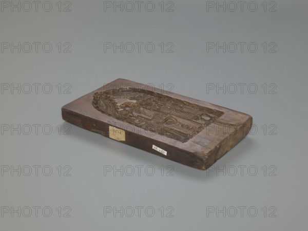 Double-sided gingerbread mould. Obverse: baptism scene; Reverse: floral ornament. Genre scenes, gingerbread moulds, folk-style decorations, baptisms, floral motifs, plant motifs, applied art, decorative art, gingerbread, figurative scenes, historical costumes. Double-sided rectangular mould: on one side a genre scene depicting a baptism — a group of figures in 17th–18th century costumes; in the centre a bearded man, probably a priest or pastor, holding a child over a baptismal font, next to him three women in long patterned dresses; the whole group surrounded by an arched window decorated with rich geometric and plant ornament. On the other side a large leaf-shaped ornament containing interwoven floral and plant motifs. 18th–19th century, Province of Silesia 1742–1918. Form: rectangular mould. Height 26 cm, width 14.5 cm, depth 2.7 cm., A rectangular wooden block with a carved recessed oval scene sits on a smooth grey background; the block shows a worn brown wood surface with darker an...