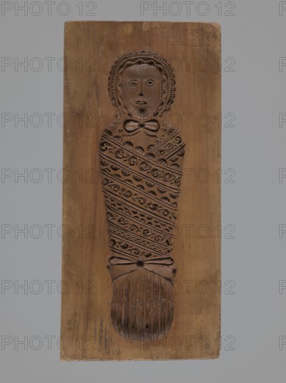Double-sided gingerbread mould. Obverse: Adam and Eve in paradise; Reverse: baby in swaddling clothes. Gingerbread mould, folk-style decorative and figurative scenes with religious motifs Adam and Eve under the Tree of Knowledge with a snake, plant and floral ornamentation. Rectangular mould, applied/decorative art. Silesia Province of Silesia, 18th–19th century. Height 22.2 cm, width 10.5 cm, depth 3.3 cm., A carved wooden plank with a relief of a wrapped figure is centered against a gray background; the wood shows brown tones with darker carved areas forming a stylized head, face, and body wrapped in patterned bands, a bow shape near the neck, and a textured rounded end, with the surrounding panel showing natural wood grain and variations in brown, and the background appearing gray.
