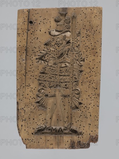 A single-sided gingerbread mold of rectangular shape with the image of a male figure in a richly decorated outfit, wearing a hat with feathers and holding a long staff; clothing characteristic of the 17th century; at the feet of the figure there is a small base on which two letters ME; gingerbread, folk-style decorations, customary scenes, staff iconographic, men, hats headgear; 18th–19th century, Province of Silesia 1742–1918; applied art; height 24.3 cm, width 14.3 cm, A rectangular wooden panel with a carved full-length figure in relief occupies the center; the wood is light brown with areas of darker brown patina and many small dark holes scattered across the surface. The carved figure wears an ornate, layered costume with detailed patterns and ruffled elements, a broad-brimmed hat with decorative plumes, and holds a long staff or spear; the carving shows textured lines and curved motifs. The bottom edge of the panel is irregular and worn with chipped and rough areas, and the backg...