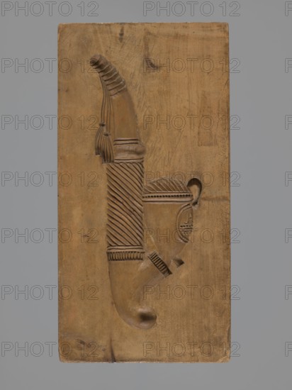 Double-sided gingerbread mould. Obverse: decorative pipe; reverse: nine fish. Double-sided rectangular mould; on one side a long decorative pipe, decorated with fluted decoration, on the stem the letter A in an oval, at the mouthpiece two decorative tassels; on the other side, nine stylized fish covered with scales, arranged in a row one next to the other. Folk-style decorative applied art; gingerbread mould, pipes, fish motifs. 18th–19th century. Province of Silesia 1742–1918. Height 22.7 cm, width 11.5 cm, depth 3.5 cm., A rectangular wooden block with a carved relief of a curved object that resembles a stylized horn or handle, carved with ridged and diagonal hatched patterns and a few tassel-like elements; the wood shows natural grain, knots, and tool marks; colors visible are brown tones in the wooden block and carving set against a uniform gray background.