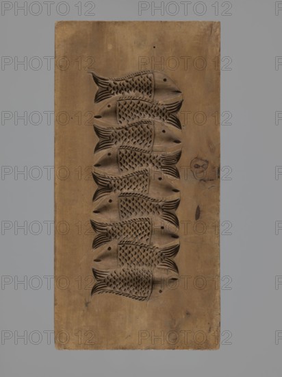 Double-sided gingerbread mould. Obverse: decorative pipe with fluted decoration; on the stem the letter A in an oval; at the mouthpiece two decorative tassels. Reverse: nine stylized fish covered with scales, arranged in a row. Folk-style decorations; applied/decorative art; gingerbread mould. 18th–19th century, Province of Silesia 1742–1918. Form rectangular; height 22.7 cm, width 11.5 cm, depth 3.5 cm., A rectangular wooden block with a vertical arrangement of a few carved fish motifs in relief running down its center, the carvings showing fish bodies with textured scale patterns and stylized fins and tails; the wood shows variations in tone and grain with knots and small imperfections, and colors visible include brown tones in the carved panel and a gray background surrounding the block.