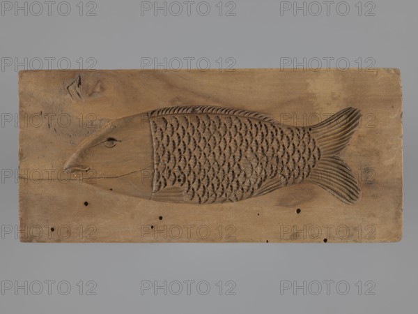 Double-sided gingerbread mold. Obverse: baby in swaddling clothes richly decorated with various geometric motifs; reverse: stylized elongated fish covered with scales. Gingerbread mold; folk-style decorations; applied/decorative art; folk customs; geometric ornaments; children; fish. 18th–19th century, Province of Silesia 1742–1918. Material: mold for gingerbread. Dimensions: height 16.7 cm; width 7.8 cm; depth 3.4 cm., A rectangular wooden panel with a carved fish motif occupies the center, the fish rendered in shallow relief with a rounded head, a carved eye and mouth, a textured body of repeating scale-shaped cutouts, and a fan-shaped tail with carved striations; the panel shows natural wood grain, knots, small holes, and areas of darker and lighter brown, set against a smooth gray background.