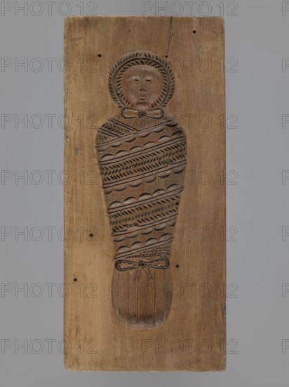 Double-sided gingerbread mold. Obverse: baby in swaddling clothes richly decorated with various geometric motifs; Reverse: stylized elongated fish covered with scales. Gingerbread mold, applied/decorative folk-style art; used for gingerbread, folk customs; geometric ornaments, children motifs. 18th–19th century, Province of Silesia 1742–1918. Material: mold. Dimensions: height 16.7 cm; width 7.8 cm; depth 3.4 cm., A rectangular wooden board in light brown with a carved relief of a wrapped figure occupies the center; the carved figure shows a rounded head with a halo-like ring and a simple facial expression, a neck tied with a bow, and a body wrapped in layers of patterned bands and scalloped edges ending in a tied section and a rounded foot-like form; the wood surface shows grain, small dark holes, and a few darker brown marks, against a plain gray background.