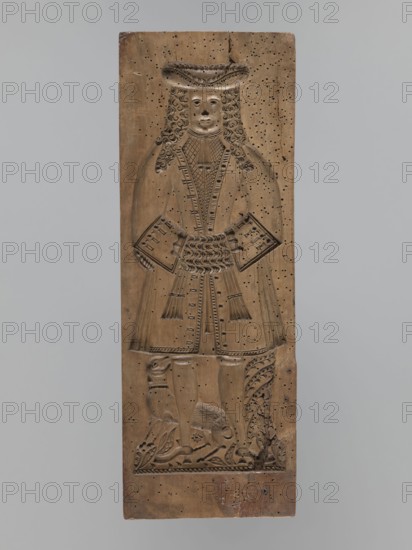 One-sided gingerbread mould with the image of a bachelor;, Rectangular mould showing a male figure standing with joined hands, wearing a patterned tricorn hat and a long wig, in a bachelor’s costume from the 2nd half of the 19th century. Attire includes a justokor, culottes, stockings and high-heeled shoes. A dog stands at the man's leg. Decorative plant motifs at the bottom. Folk-style decorations; applied and decorative art. Materials: gingerbread mould. Date: 17th century or early 18th century; also associated with 18th–19th century. Region: Silesia Province of Silesia, 1742–1918. Dimensions: height 50 cm, width 18 cm, depth 4.2 cm., A tall vertical wooden panel carved in a warm brown tone shows a full-length figure of a person in period dress with long curly hair and a wide brimmed hat, hands held at the waist holding rectangular objects, and a long coat with decorative carved patterns; the lower portion of the panel features carved legs and shoes above a carved ground with floral ...