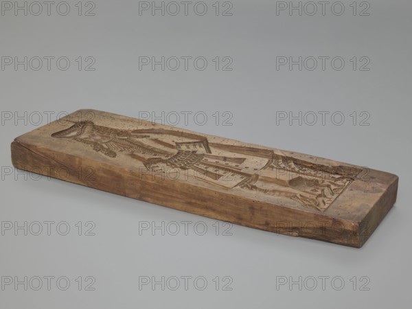 One-sided gingerbread mould with the image of a bachelor. One-sided rectangular mould showing a male figure standing with joined hands, wearing a patterned tricorn hat and a long wig, in a bachelor’s costume from the 2nd half of the 19th century; alternatively dated 17th century or early 18th century, dressed in a justokor, culottes, stockings and high-heeled shoes. A dog at the man's leg and decorative plant motifs at the bottom. Applied/decorative folk-style gingerbread mould, plant decorations and motifs, dogs, male figure. 18th–19th century, Province of Silesia 1742–1918. Form dimensions: height 50 cm, width 18 cm, depth 4.2 cm., A rectangular wooden block rests flat on a smooth surface, showing a carved recessed design on its top face; the block displays a range of brown wood tones from light tan to deeper chestnut with visible grain and some darker knots and spots, and the carved area reveals varying depths and shadows in similar brown hues. The carving depicts a stylized humanli...