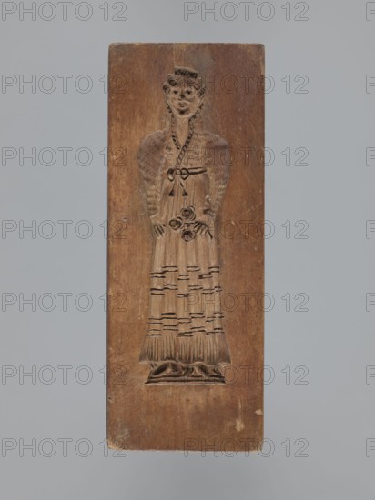Double-sided gingerbread mould. Obverse: female figure in a long, patterned, richly decorated dress, wearing a hat decorated with an egretta. Reverse: male figure wearing a fur hat and a fur-lined coat over an embroidered shirt, holding a long pipe. Plant decoration at the bottom of both figures. Woodcarving, folk-style decorations; gingerbread moulds; decorative/applied art; historical costumes. 18th–19th century, Province of Silesia 1742–1918. Rectangular form; height 19 cm, width 9 cm, depth 3.3 cm., A rectangular wooden block carved in relief with the figure of a standing person wearing a long dress and a headpiece; the wood shows a warm brown tone with lighter and darker variations and areas of wear, and the background around the carving is a similar brown; the carved figure has textured details on the sleeves and skirt, a belt with a tied bow, and floral-like shapes held near the waist, with carved folds at the hem and a small raised base beneath the feet; the overall scene is se...