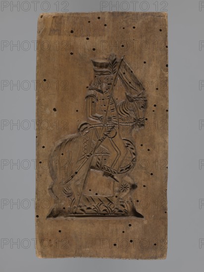 Double-sided gingerbread mould with the image of a rider on both sides, gingerbread; subject: horses, riders in historical military costumes hussar-type and another military uniform, sabres, pole weapon lance, horse head with decorative covering; style: folk-style decorations, plant motifs; materials/function: gingerbread mould, decorative art; form: double-sided, rectangular mould; inscriptions/details: one side—rider in military uniform decorated with braiding, high mirliton-type headgear, holding a sabre, plant decoration at the bottom; other side—rider in a different military uniform wearing a cap with a feather, holding a pole weapon, sabre at his side, horse head decorated, decorative plant motifs at the bottom; date: 18th–19th century; place: Province of Silesia 1742–1918; dimensions: height 17.2 cm, width 9 cm, depth 2 cm., A rectangular wooden panel with a carved relief of a mounted figure occupies the center; the wood is brown with a weathered surface and shows a pattern of s...