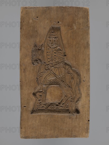 Double-sided gingerbread mould with the image of a rider on both sides;, Gingerbread; folk-style and plant decorations; horses, uniforms, sabres, plant motifs; hussars; horse rider iconography; historical costumes. Rectangular mould: one side shows a rider in a military uniform decorated with braiding, likely a hussar, wearing a high mirliton-type headdress and holding a sabre, with plant decoration at the bottom. The other side depicts a rider in a different military uniform, wearing a cap with a feather, holding a pole weapon lance, with a sabre at his side and the horse’s head covered decoratively; decorative plant motifs at the bottom. 18th–19th century, Province of Silesia 1742–1918. Form: double-sided. Height 17.2 cm; width 9 cm; depth 2 cm., A rectangular wooden panel with a carved relief of a mounted figure occupies the center. The panel is brown with lighter and darker wood grain variations. The carved scene shows a person seated on a horse; the person wears a pointed headpiec...