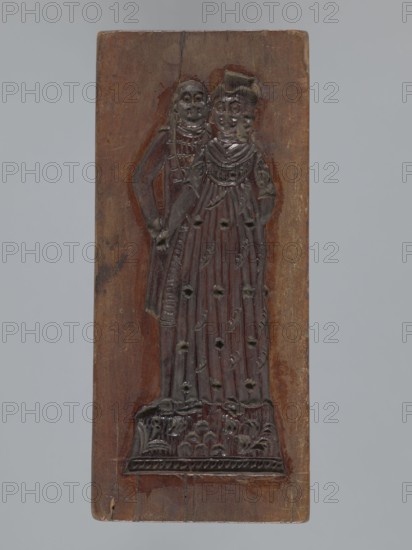 Double-sided gingerbread mold. Obverse: couple holding hands—woman in a richly decorated long dress with a hat, man in a caftan and short wig; costumes typical of the 18th century; decoration of plant elements at the bottom. Reverse: three empty rectangles. Woodcarving; gingerbread mold; genre scene of a couple in love, female and male figures, plant motifs. 18th–19th century, Province of Silesia 1742–1918. Applied art. Height 20 cm; width 8.7 cm., A rectangular wooden panel with a carved relief of two standing figures occupying most of its central area, the wood showing brown and darker brown tones with some lighter brown wear; the carved figures are darker, almost blackish-brown, with one figure slightly behind the other and both wearing long, draped garments with vertical folds and decorative impressions; the front figure appears to have a rounded hairstyle or head covering and a high neckline, while the rear figure shows a suggestion of layered clothing at the shoulders; the base o...