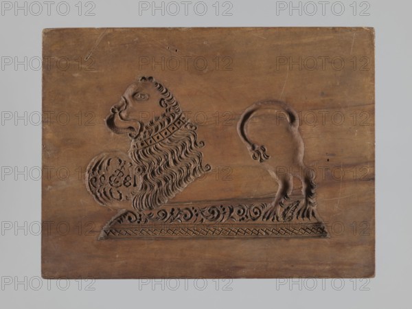 Double-sided gingerbread mould. Rectangular wooden form, height 25.8 cm, width 19.4 cm, depth 4.3 cm. One side: stylised lion with abundant mane standing on a base decorated with plant-twig ornament, holding a ball engraved with the date 1829 and a symbol of an anchor intertwined with the letter S; floral and plant decoration at the bottom. Reverse: rider on horseback in early 19th-century riding attire jacket, breeches, top hat, holding a riding crop; at the horse's front legs a round shield engraved 1831 and the same anchor-and-S symbol mark of the woodcarver or owner of the form; floral and plant ornament at the bottom. Materials/function: applied art; gingerbread mould. Iconography: figurative scenes, lions, horse, historical costumes, plant motifs. Region/date: Province of Silesia 1742–1918; dates on mould: 1829 and 1831. Creator: unknown., A rectangular wooden plaque with a carved relief of a stylized lion in profile, showing its head, mane, front paw resting on a rounded object,...