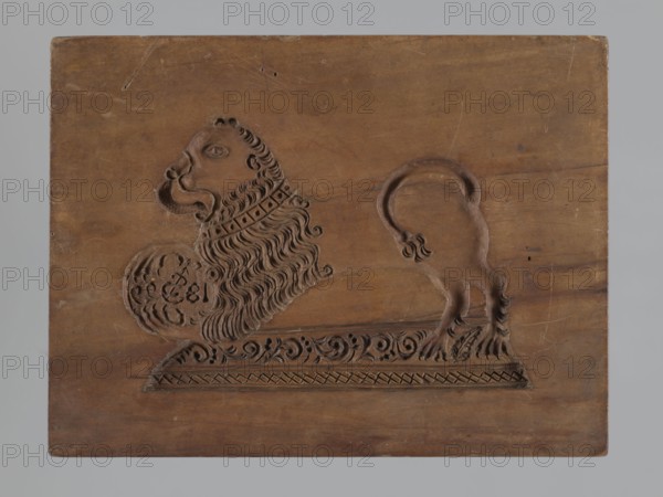 Double-sided gingerbread mould. Rectangular form, height 25.8 cm, width 19.4 cm, depth 4.3 cm. Obverse: image of a rider — figure of a cavalier on horseback in early 19th-century riding attire jacket, breeches, top hat, holding a riding crop; at the horse’s front legs a round shield engraved “1831” and a mark showing an anchor intertwined with the letter “S”; bottom decorated with floral and plant ornament. Reverse: stylised lion with abundant mane standing on a base decorated with plant-twig ornament, holding a ball engraved “1829” and the same anchor-with-S mark; bottom with floral and plant decoration. Materials/technique: carved mould for gingerbread applied art. Iconography: rider on a horse, lions, plant motifs; historical costumes. Place/date attribution: Silesia, Province of Silesia 1742–1918; dates on images 1829 and 1831., A carved wooden panel shows a stylized animal with a large mane and a curled tail, rendered in relief against a plain wooden background; the carving includ...