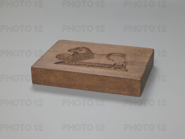 Double-sided gingerbread mould. Rectangular form, wood; height 25.8 cm, width 19.4 cm, depth 4.3 cm. One side: stylised lion with abundant mane standing on a base decorated with plant-twig ornament, holding a ball engraved with the date 1829 and a mark of an anchor intertwined with the letter S; floral and plant decoration at the bottom. Reverse: rider cavalier on horseback in early 19th-century riding attire jacket, breeches, top hat, holding a riding crop; a round shield at the horse's front legs engraved 1831 and the same anchor-and-S mark likely the woodcarver's or owner's mark; floral and plant ornament at the bottom. Iconography: rider on a horse, lions, figurative scenes, historical costumes, plant motifs. Gingerbread mould; applied art. Province of Silesia 1742–1918., A rectangular wooden block with a carved relief on its top surface depicting a stylized animal figure with a mane and curled tail, placed on a smooth light gray surface. The wood shows warm brown tones with variat...