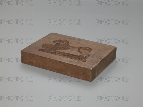 Double-sided gingerbread mould. Rectangular shape, height 25.8 cm, width 19.4 cm, depth 4.3 cm. Obverse: image of a rider on horseback in early 19th-century riding attire jacket, breeches, top hat, holding a riding crop; at the horse’s front legs a round shield engraved 1831 and a symbol of an anchor intertwined with the letter S mark of the woodcarver or owner; bottom floral and plant ornament. Reverse: stylised lion with abundant mane standing on a base decorated with plant twigs, holding a ball engraved 1829 and the same anchor-and-S symbol; bottom floral and plant decoration. Materials/genre: applied art, gingerbread mould; motifs: figurative scenes, lion, horse, historical costume, plant motifs; region: Silesia Province of Silesia, 1742–1918., A rectangular wooden block with a carved relief on its top surface sits against a plain light-gray background, the block showing warm brown and tan wood tones and a polished finish, with visible grain and a few darker brown patches and subtl...