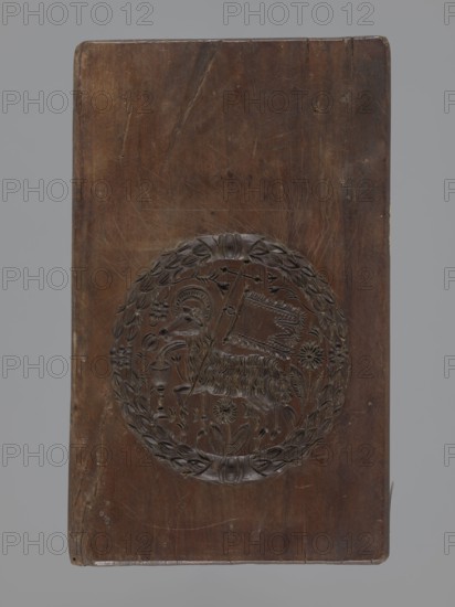 Double-sided gingerbread mould. Obverse: Easter Lamb; reverse: lady at a spinning wheel and three triangles. Woodcarving, gingerbread mould, applied decorative art. Icons: Lamb of God holding a banner with halo, chalice with blood flowing into it; background strewn with numerous flowers; surrounded by a laurel wreath. Other side: female figure seated at a spinning wheel in a long decorative dress with a deep neckline, plus three triangular motifs decorated with geometric elements. Floral and geometric motifs, laurel wreath ornament. Double-sided, rectangular mould, four motifs on one side. 18th–19th century, Province of Silesia 1742–1918. Height 28 cm; width 14.4 cm; depth 2.5 cm., A rectangular wooden board with a carved circular medallion near its center; the board shows a worn, smooth surface with visible scratches, small dents, and darker and lighter brown tones throughout, and the carved medallion features a raised wreath-like border and an intricate central relief depicting an an...