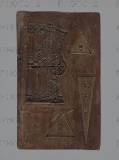 Double-sided gingerbread mould. Obverse: Easter Lamb holding a banner, haloed, with a chalice in front catching flowing blood; background strewn with numerous flowers and surrounded by a laurel-leaf wreath. Reverse: female figure seated at a spinning wheel in a long decorative dress with a deep neckline, accompanied by three triangular motifs decorated with geometric elements; four motifs on this side. Woodcarving; gingerbread mould; decorative and applied art; religious and festive iconography. 18th–19th century, Province of Silesia 1742–1918. Rectangular form, double-sided; height 28 cm, width 14.4 cm, depth 2.5 cm., A rectangular wooden panel with a rich brown color and visible wood grain, featuring carved designs including a figure of a seated person with detailed clothing and a wheel beside them, decorative triangular and elongated wedge-shaped motifs, and various carved circular elements; the panel shows surface wear, scratches, small dents, and slightly rounded corners against a...