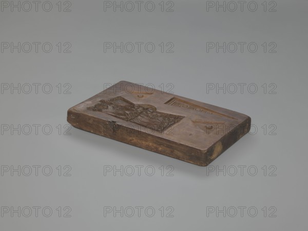 Double-sided gingerbread mould. Obverse: Easter Lamb holding a banner, haloed, with a chalice collecting flowing blood, background strewn with numerous flowers, surrounded by a wreath woven from laurel leaves. Reverse: female figure seated at a spinning wheel in a long decorative dress with a deep neckline, plus three triangular motifs decorated with geometric elements. Rectangular mould, four motifs on one side. Material: wood woodcarving; function: gingerbread mould. Style/subjects: religious motifs Lamb of God, festive traditions, spinning wheel, female figure, laurel wreath, floral and geometric ornament. Date: 18th–19th century. Region: Province of Silesia 1742–1918. Dimensions: height 28 cm, width 14.4 cm, depth 2.5 cm., A rectangular carved wooden block rests on a smooth pale gray surface against a lighter gray background; the block is a warm brown with darker brown areas and some lighter tan spots at the edges, and its top surface features recessed carved shapes including a rec...