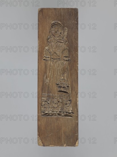 Double-sided gingerbread mold. Obverse: female figure; Reverse: baby in swaddling clothes. Woodcarving; gingerbread mold for decorative/applied art. Single-sided rectangular mold with image of a female figure in Silesian folk costume—bonnet, large bow tied under the neck, long dress with short puffed sleeves—dress decorated at the bottom with a floral and plant ornament; figure holds a basket with flowers in her left hand. Other side shows a baby in decorative swaddling clothes. Folk-style decorations, floral motifs. 18th–19th century, Province of Silesia 1742–1918., A vertical wooden panel carved in brown tones shows a relief of a standing figure wearing a dress with puffed sleeves and a large bow at the neck, the figure holding a small rounded object near the waist and standing above a decorative carved base with floral motifs; the wood surface shows lighter and darker brown variations and areas of wear at the edges against a neutral pale gray background.
