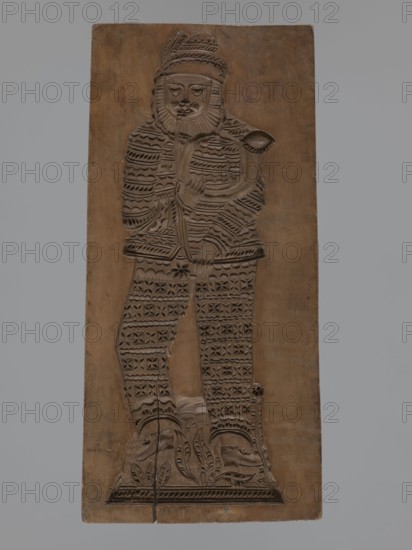 Double-sided gingerbread mould. Obverse: figure of a bearded man playing a horn; reverse: several small elements including houses with tiled roofs, a cradle decorated with a flower motif, and a chest or bed decorated with a plant twig. Folk-style decorations and figurative scenes with geometric and plant ornaments; motifs include horn musical instrument, cradles, houses, male figure. Double-sided rectangular form richly decorated with various geometric motifs; man wears patterned clothes and a hat with a feather. 18th–19th century, Silesia Province 1742–1918. Materials: form for gingerbread applied/decorative art. Height 30 cm, width 13.8 cm, depth 2 cm., A rectangular wooden panel carved in brown tones shows a full-length stylized figure in low relief wearing a patterned outfit and a hat, holding a curved object near the chest; the figure has a carved beard and facial features with closed eyes, and the clothing and legs are decorated with repeating geometric and floral motifs; the bas...